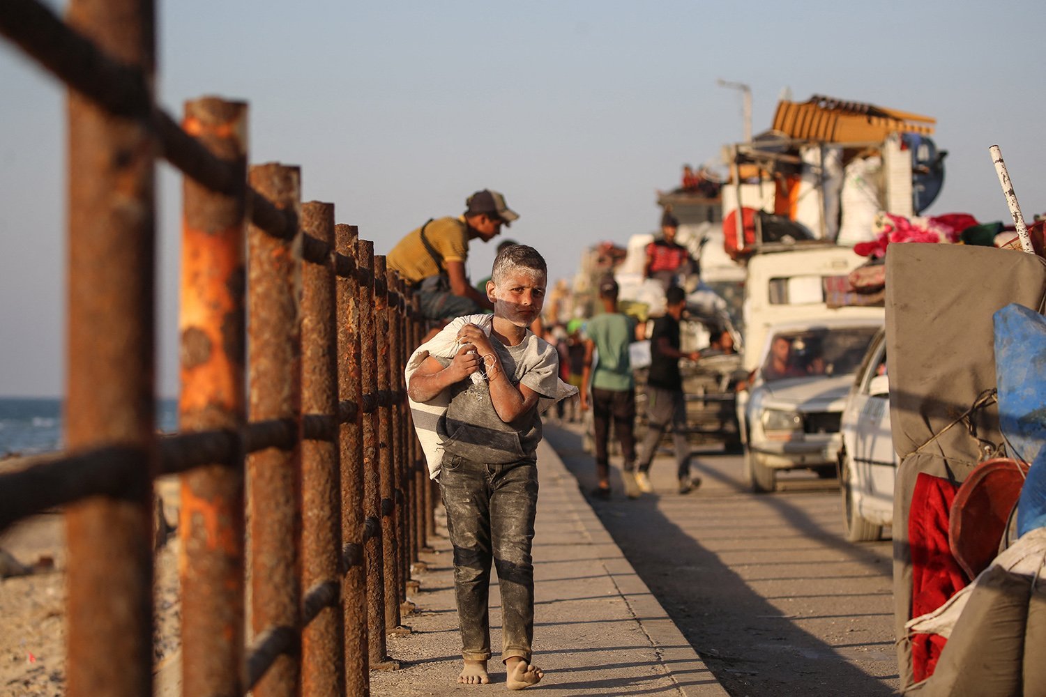 A child carrying his luggage walks on the sidewalk next to the fence. In the distance, cars loaded with people and property appear.