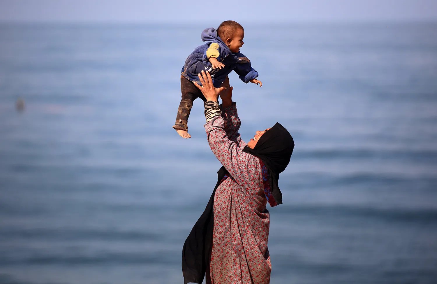 A veiled woman throws a child into the air, and they both smile. Behind them is the sea.