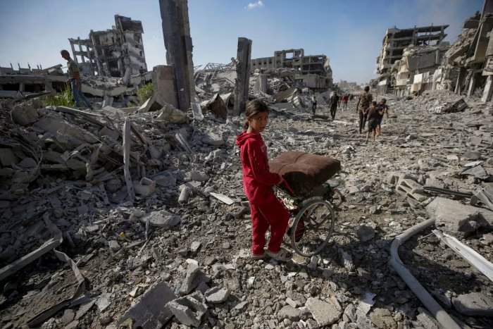 A girl wearing a red tracksuit walks through rubble in Gaza