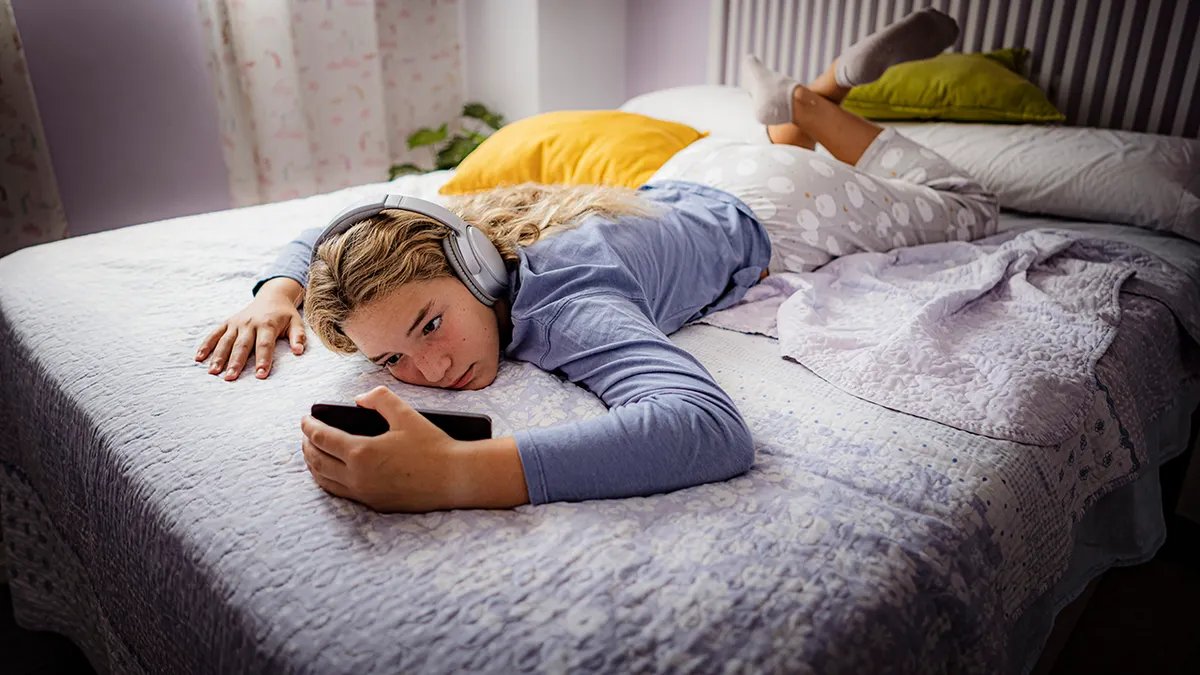 Teenage girl lying on bed looking sad while watching something on her phone, wearing big headphones.