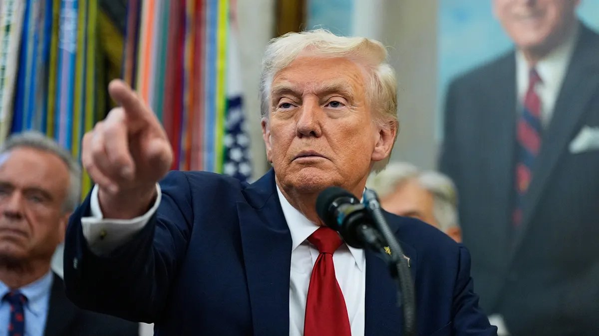 President Donald Trump wears a blue suit and red tie as he gestures to a reporter in the Oval Office.