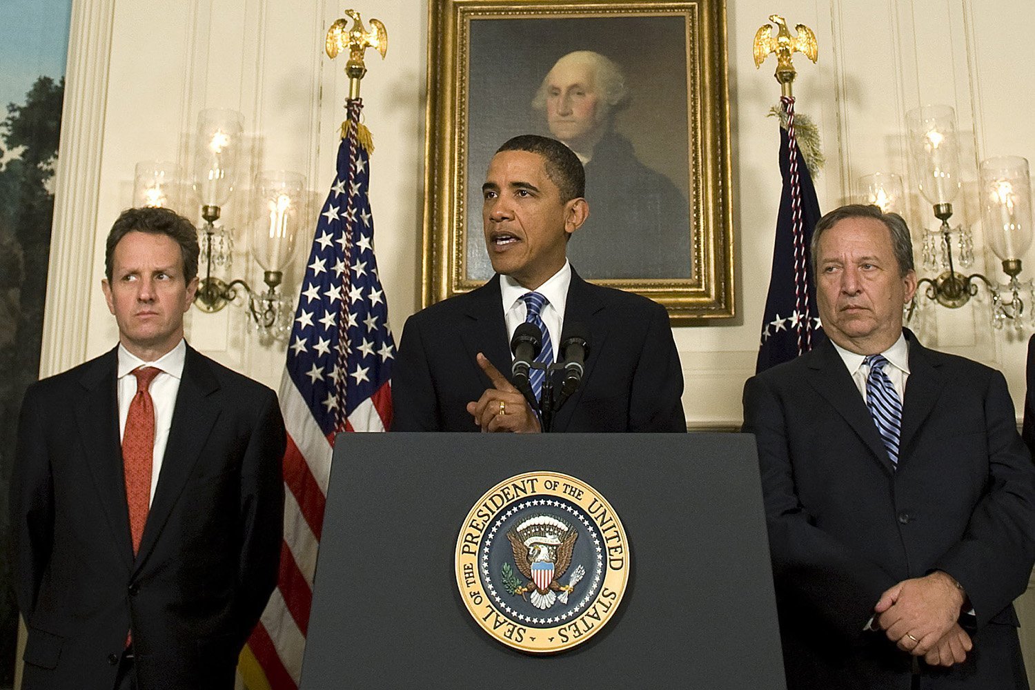 Barack Obama, dressed in a black suit and tie while standing at a podium affixed with the presidential seal, speaks between two men standing on either side. Both of the other men wear dark suits and ties. A portrait of George Washington hangs on the wall behind them.