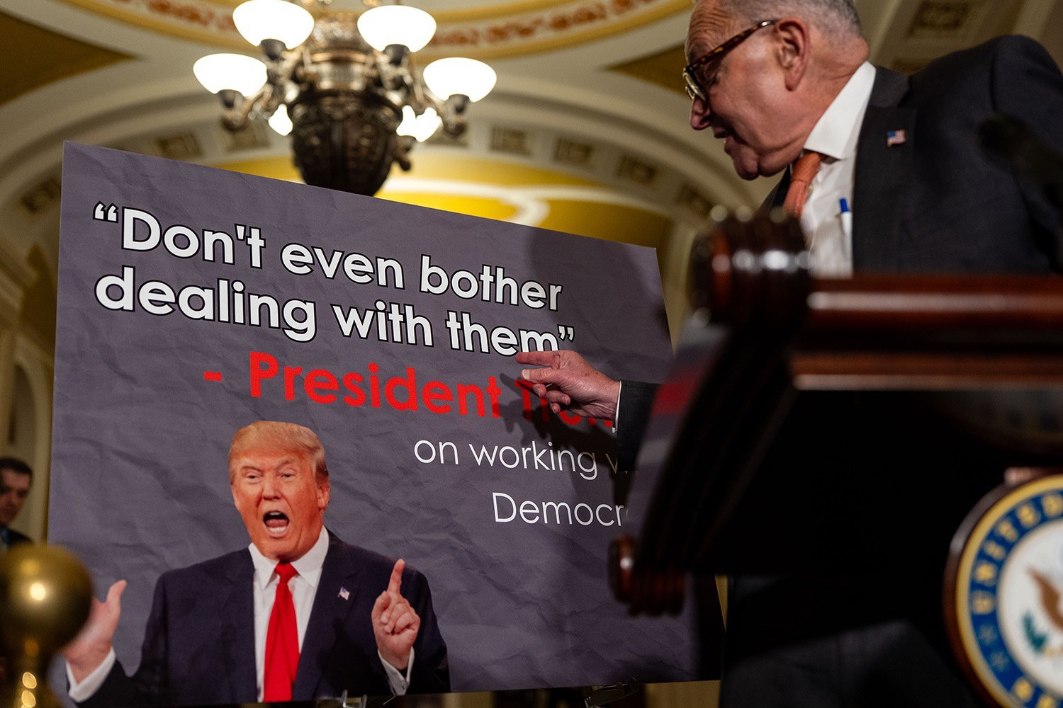 Chuck Schumer is seen in profile as he gestures to a sign that reads "Don’t even bother dealing with them. -President Trump on working with Democrats" with a photo of Trump printed beside the words.