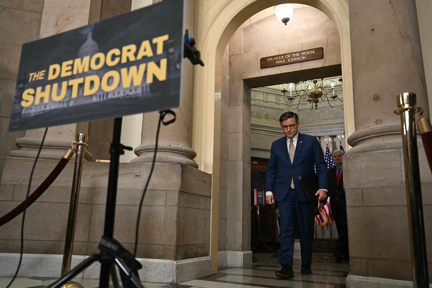 Mike Johnson walks through a large arched doorway as he arrives to a news conference, with a roped off area behind a sign that reads "The Democrat Shutdown."