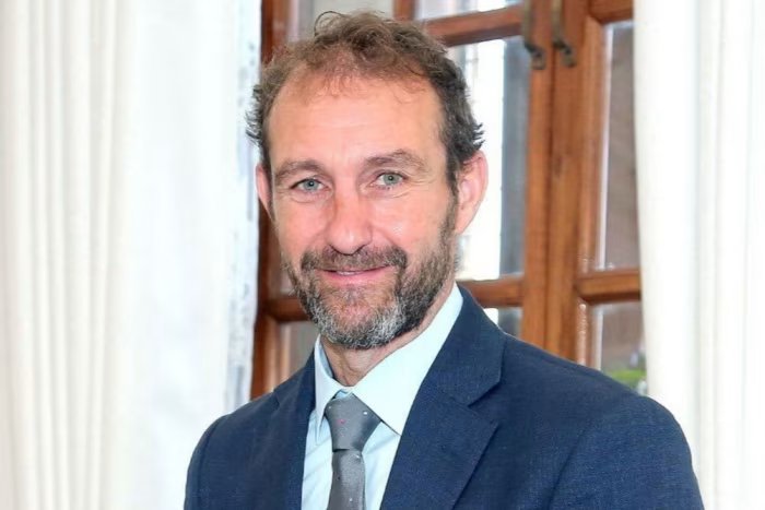 Matt Collins wears a blue suit and smiles in front of a window with white curtains.