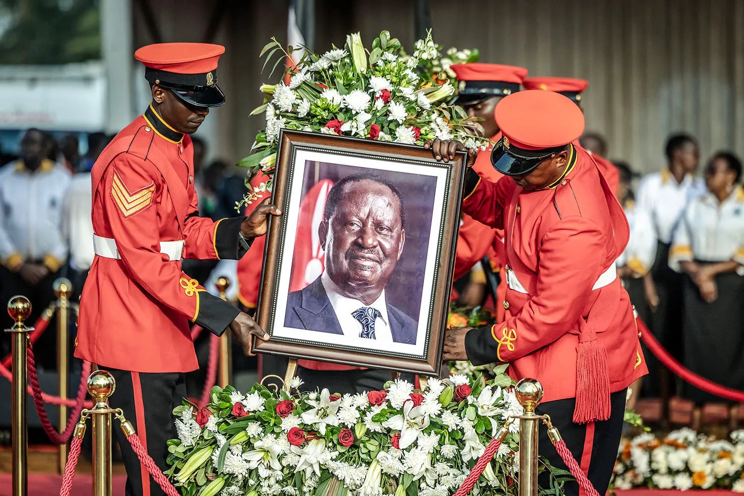 Two members of the Kenya Army, dressed in formal ceremonial uniforms with red jackets and red hats, place a framed portrait of Raila Odinga in front of a wreath of flowers surrounded by a red and gold stanchion barrier.