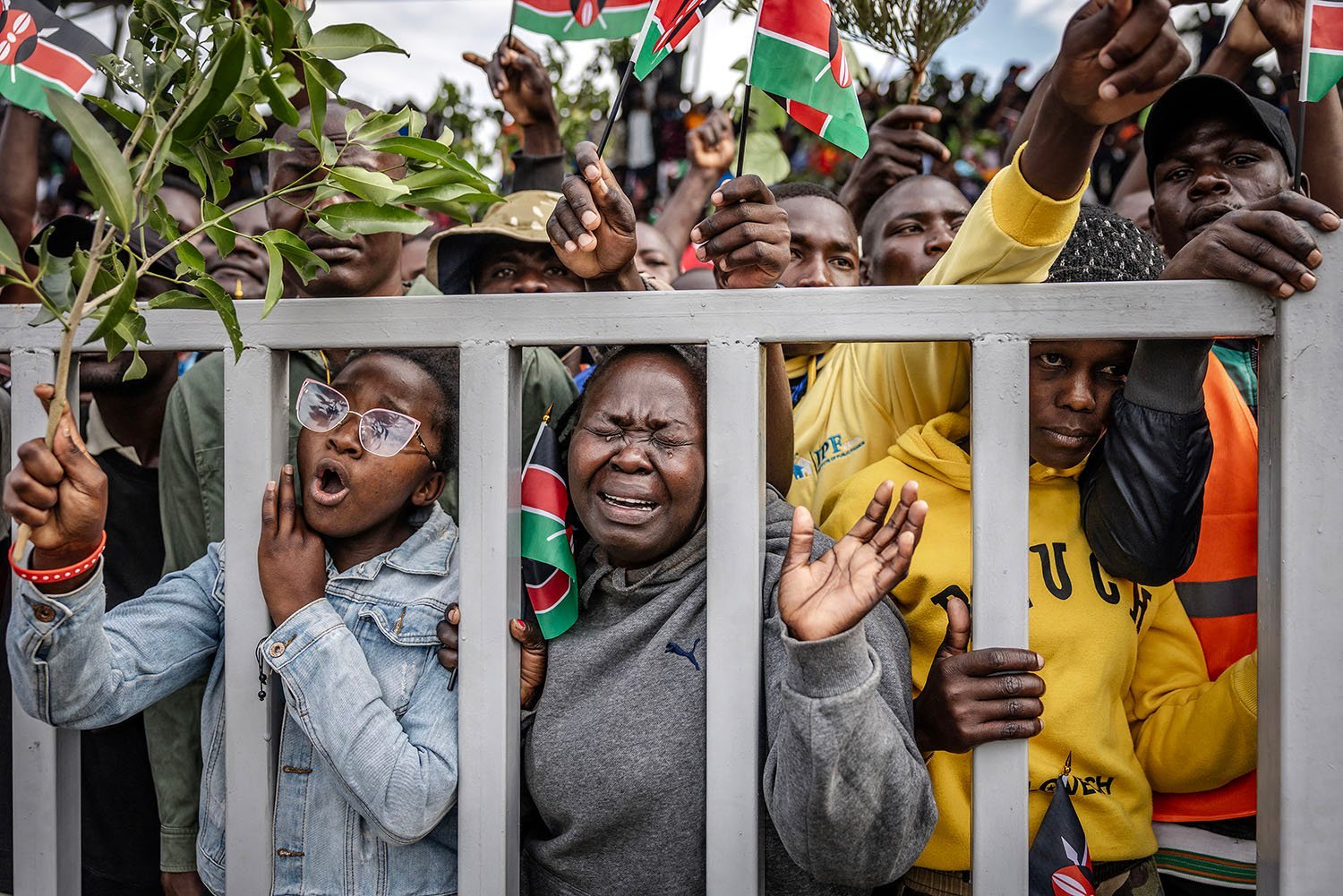 A few people lean against a white fence at the front of a bigger crowd of dozens of people, who hold up tree branches and small Kenyan flags. One woman in the front and center cries; the woman to her right shouts.