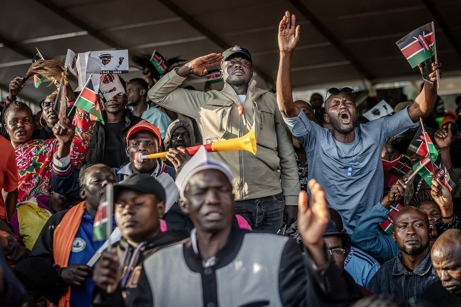 Amid a crowd of people gathered in stadium seats and waving Kenyan flags and their arms, a man stands in a salute, gazing ahead with a solemn expression.
