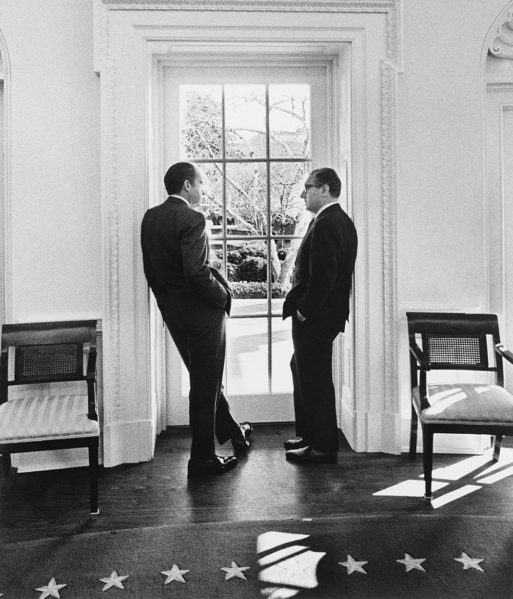 Two men lean in the frame of a tall window flanked by arm chairs.
