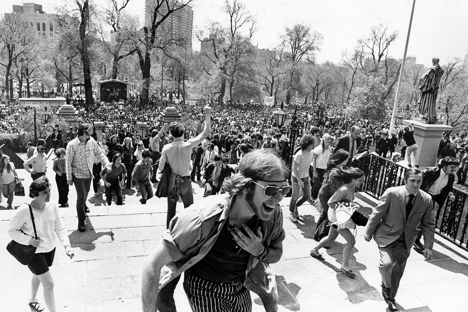 A man with long hair and sunglasses moves toward the camera as a crowd of protesters is seen behind him.