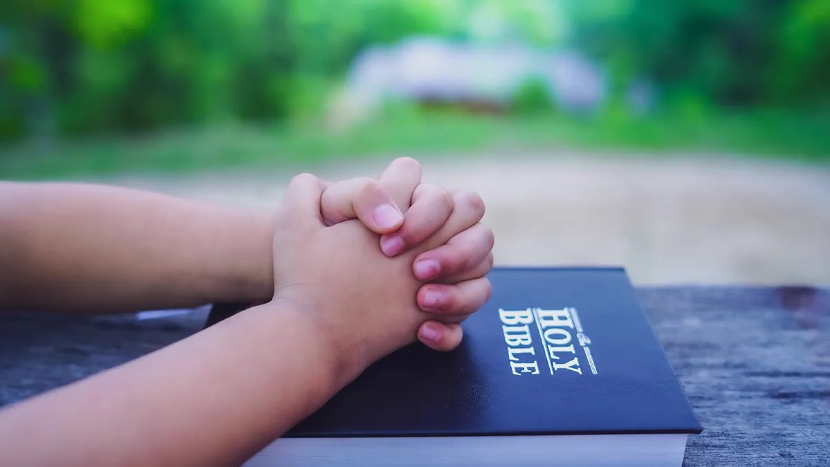 Close-up of a child's hands folded in prayer, leaning on the Bible