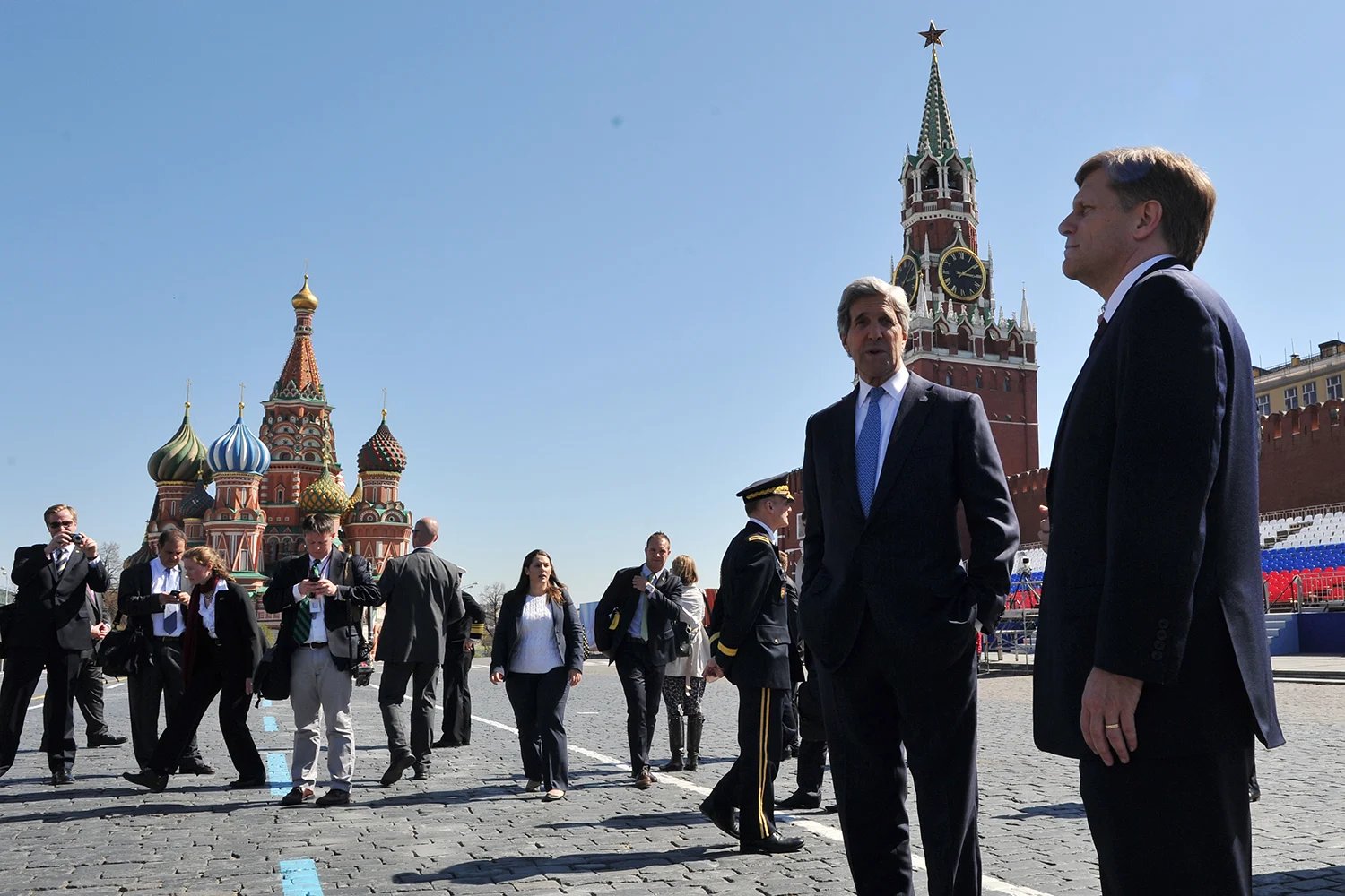 Former U.S. Secretary of State John Kerry and McFaul, dressed in suits, stand and speak to each other in the Red Square in Moscow beneath a cloudless blue sky. A group of photographers stand in the background and take photos of them. Behind them rise the colorful domes of the Kremlin.