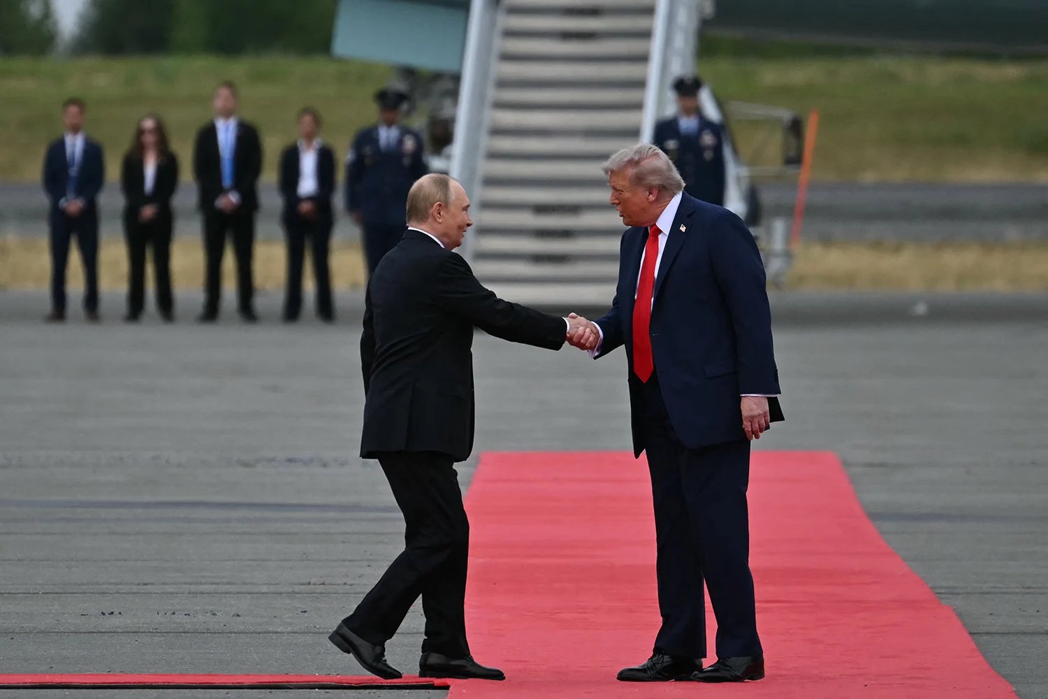 Trump and Putin are seen in profile as they shake hands while standing on a red carpet on the tarmac of an airport. The stairs to a aircraft are visible behind them.