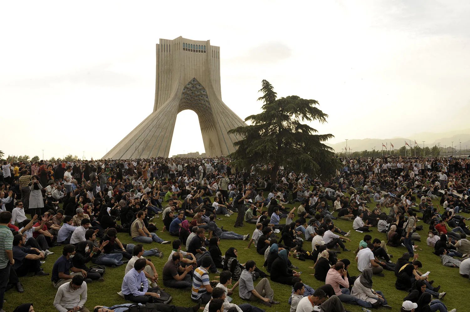 People sit scattered on a vast lawn in front of a large arched monument.