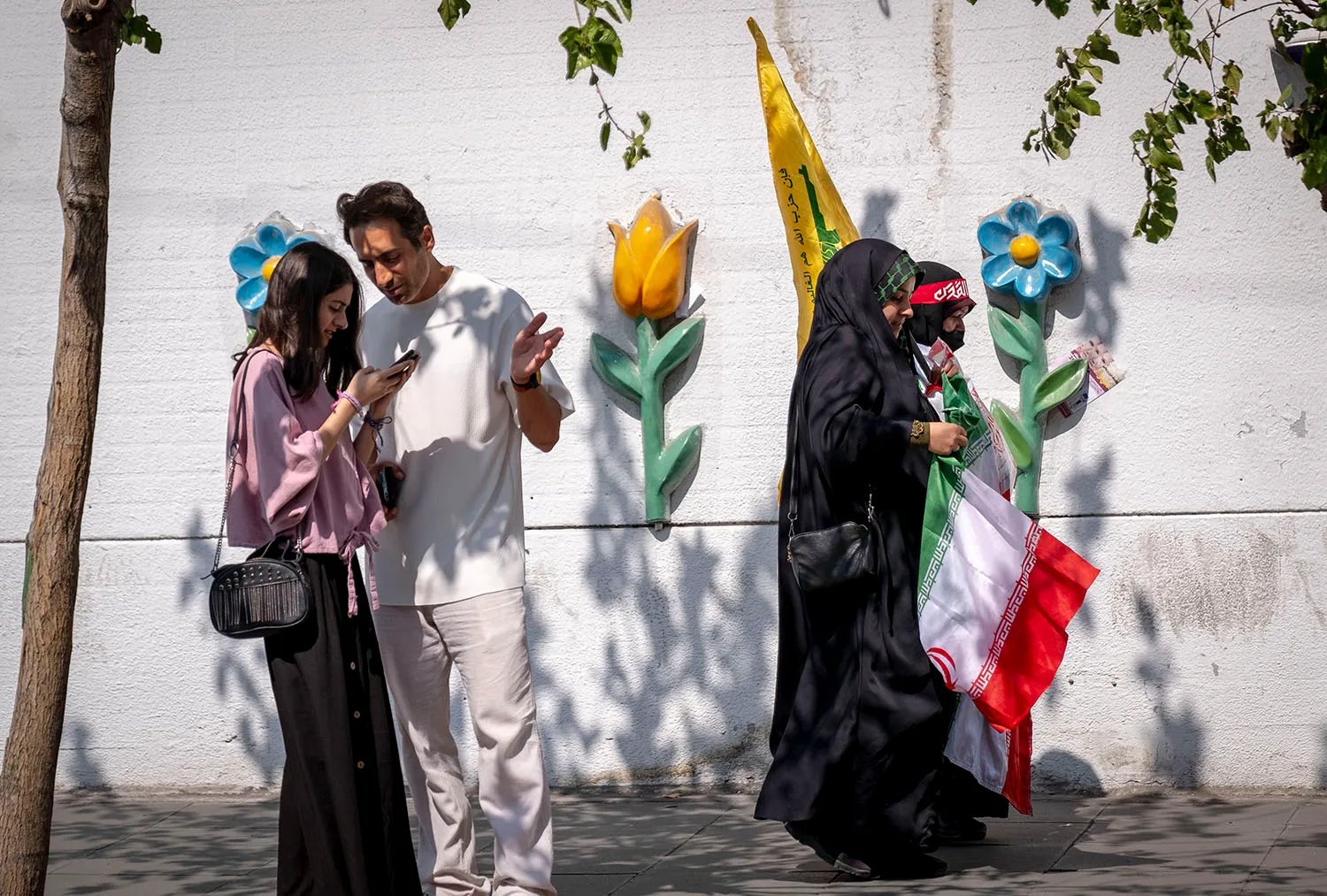 Two women in veils and head coverings carry flags as they walk by a flower-decal-covered wall. A young man in stand at left looking at a phone.