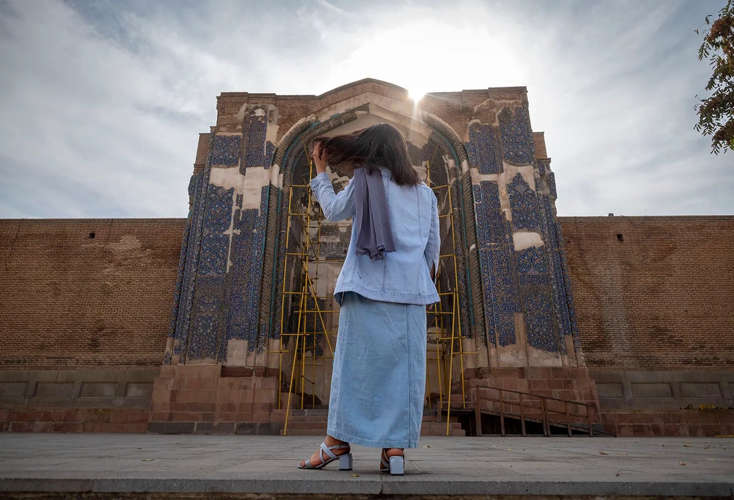 A woman with her hair loose and a scarf around her neck stands facing a mosque with the sun glinting behind it.
