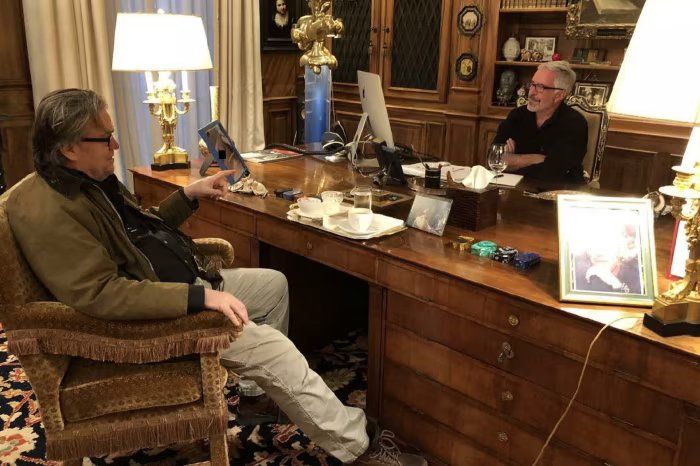 Two men sit across from each other in a wood-paneled office, one sitting at a desk and the other in an armchair holding something. The desk contains drinks, framed pictures, and stationery.