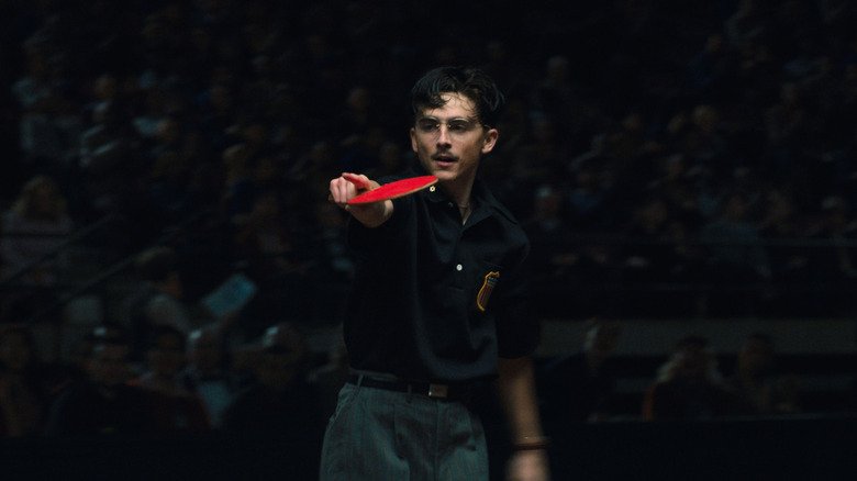 Timothée Chalamet as Marty Mouser holds a table tennis paddle and points in front of a crowd of people in Marty Supreme