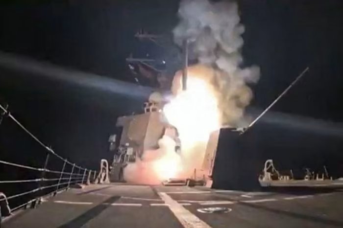 A missile is launched from a US Navy ship at night, with flames and smoke visible around the missile launch point.