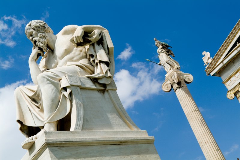 Marble statues of Socrates seated in thought and a standing Athena atop a tall column, set against a bright blue sky.