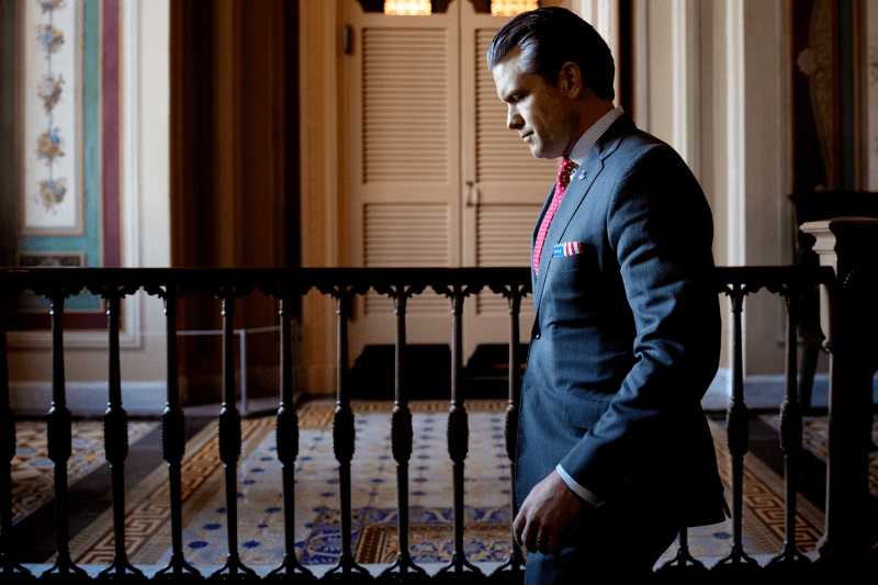 Hegseth is seen in profile as he walks through a hallway on Capitol Hill. He wears a dark suit and red tie, and his face his partly in shadow.