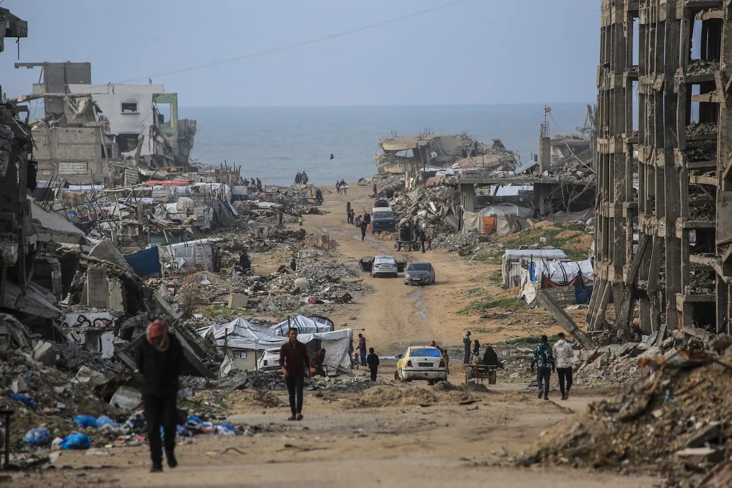 Collapsed and partially-destroyed buildings line a dirt road that leads toward the sea. Rubble, broken concrete, and makeshift shelters line the sides of the street. Several people walk along the road.