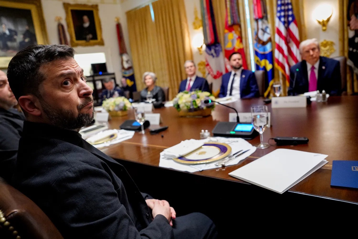 Ukrainian President Volodymyr Zelensky, dressed in dark clothing, sits at a large conference table in an ornate government meeting room, looking over his shoulder toward the camera. Several other officials, including Trump, sit around the table in the background, with microphones, documents, water glasses, and place settings visible. U.S. flags, military banners, framed portraits, and gold curtains decorate the room.