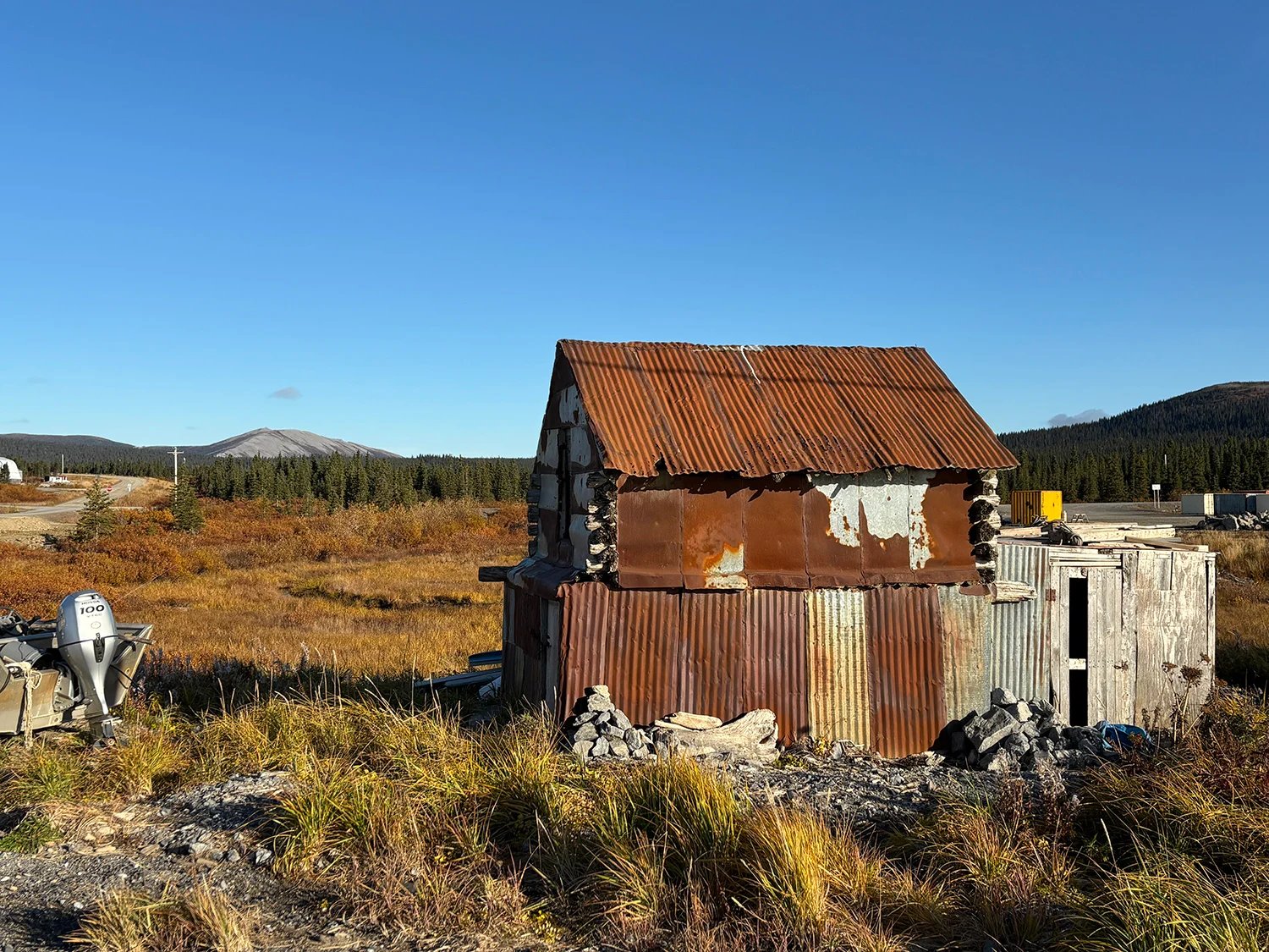 A dilapidated building with a rusty corrugated roof in a field against a blue sky.
