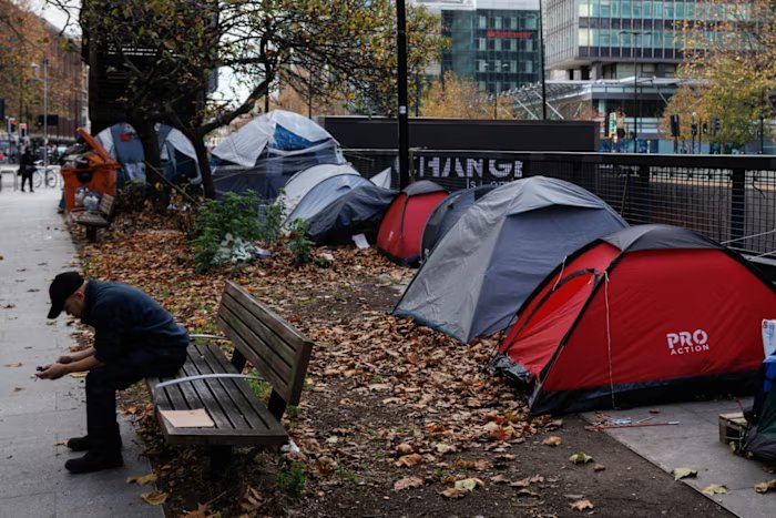 Tents were set up along a leaf-strewn sidewalk next to a busy road. A person sits on a nearby bench and looks down.