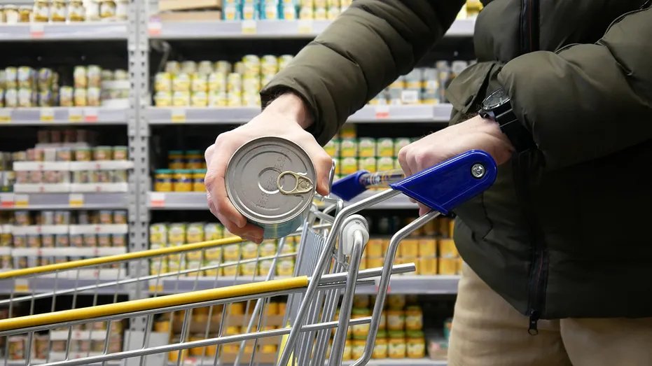 Close-up of a grocery store shopping cart and a male buyer placing a box of canned foods in it
