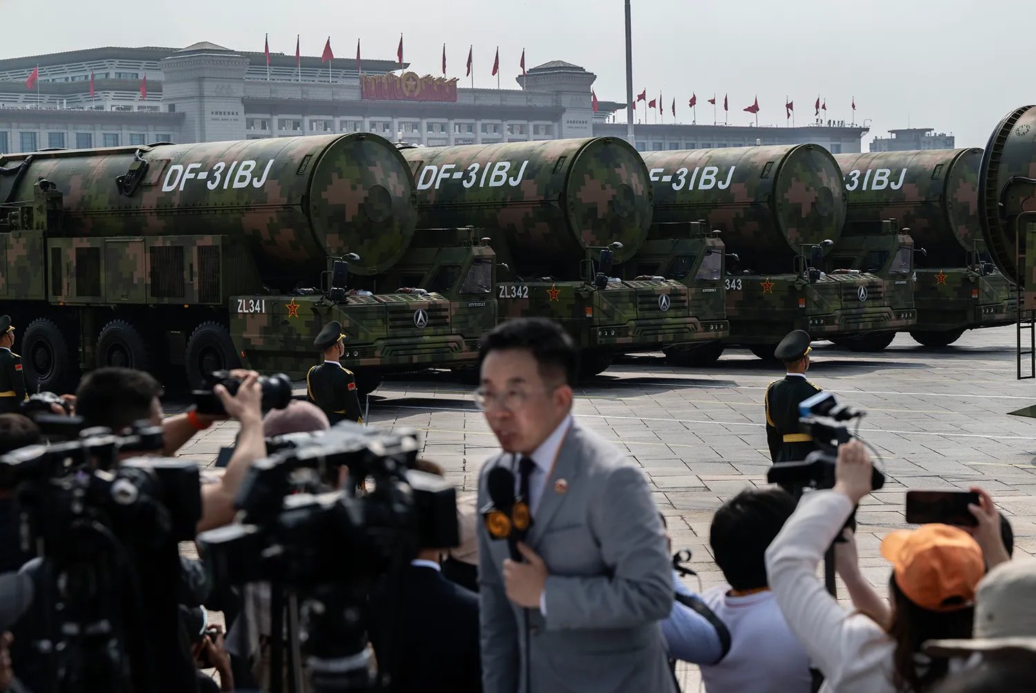 A news reporter holding a microphone is surrounded by cameras as he stands in front of a row of large military equipment.