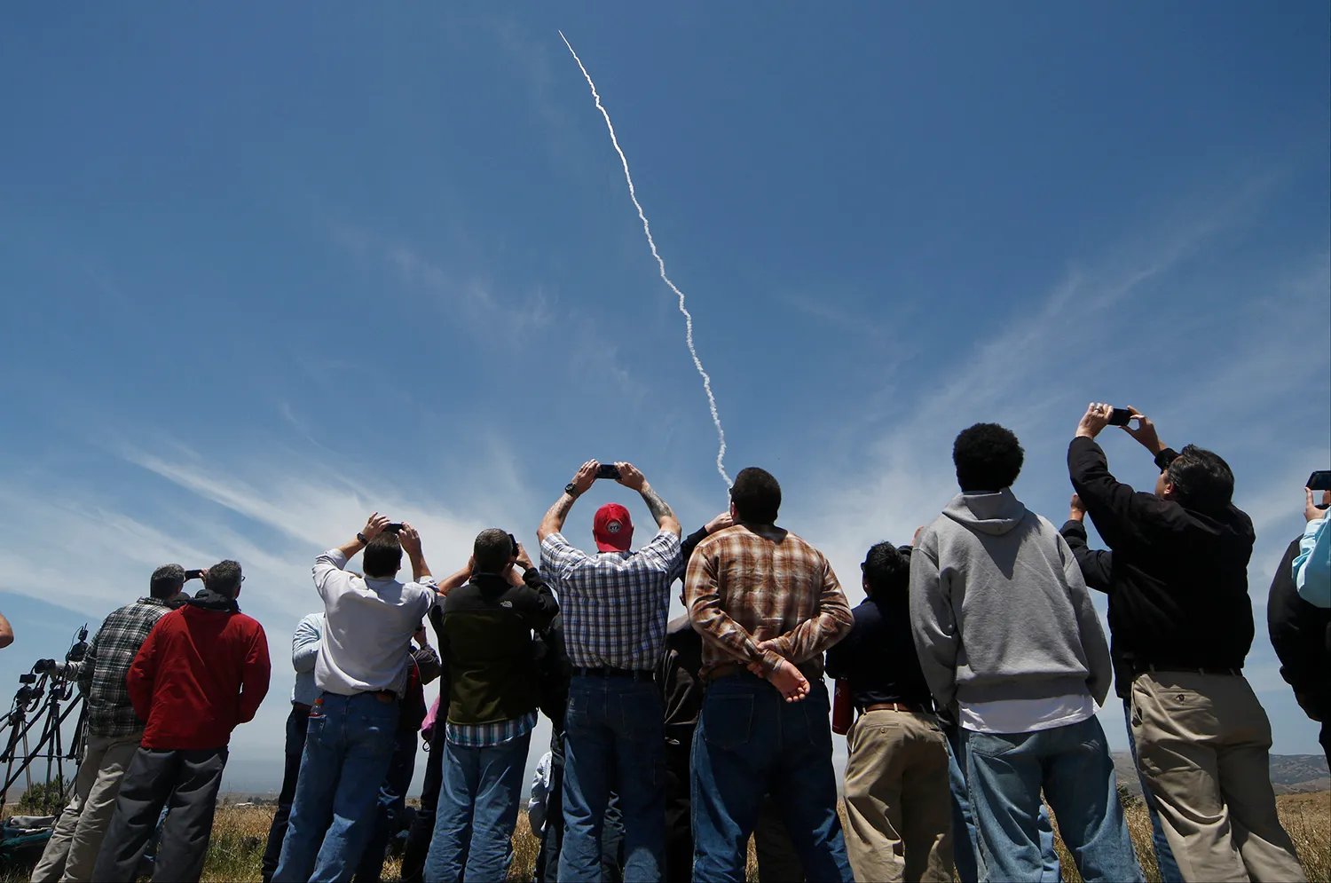 People, one holding a cell phone to record, look up at the sky as a thin line of white from a missile arcs upward.