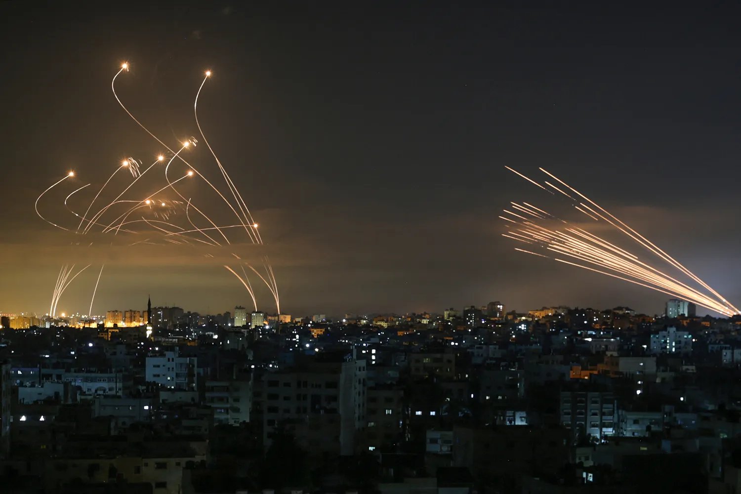 Missiles create arcs of light in the night sky with city lights and a landscape below.