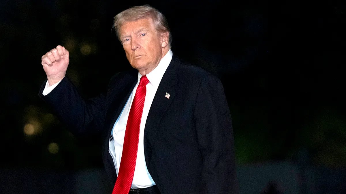 President Donald Trump wears a dark suit and red tie as he walks with his arm raised in a gesture.
