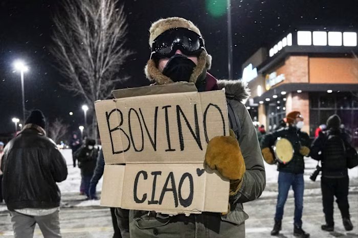 A protester wearing winter clothes holds a cardboard sign with the words: "Bovino Chow" During a night demonstration.